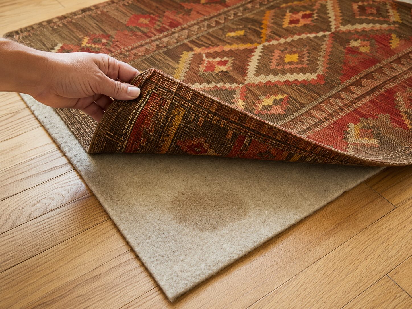 Hand lifting corner of vintage Turkish kilim rug to reveal rug pad underneath on oak hardwood floor — checking for hidden pet odor source — Kirmen Rugs