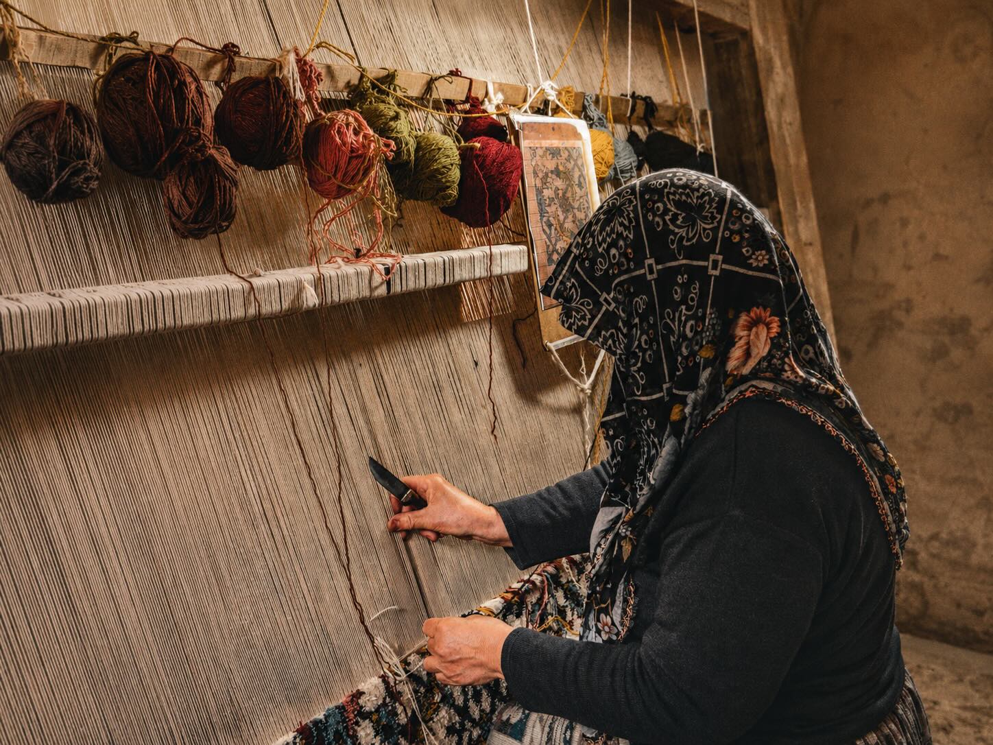 A traditional weaver in Cappadocia hand-knotting a rug on a vertical loom, with colorful wool yarns and a pattern chart hanging above