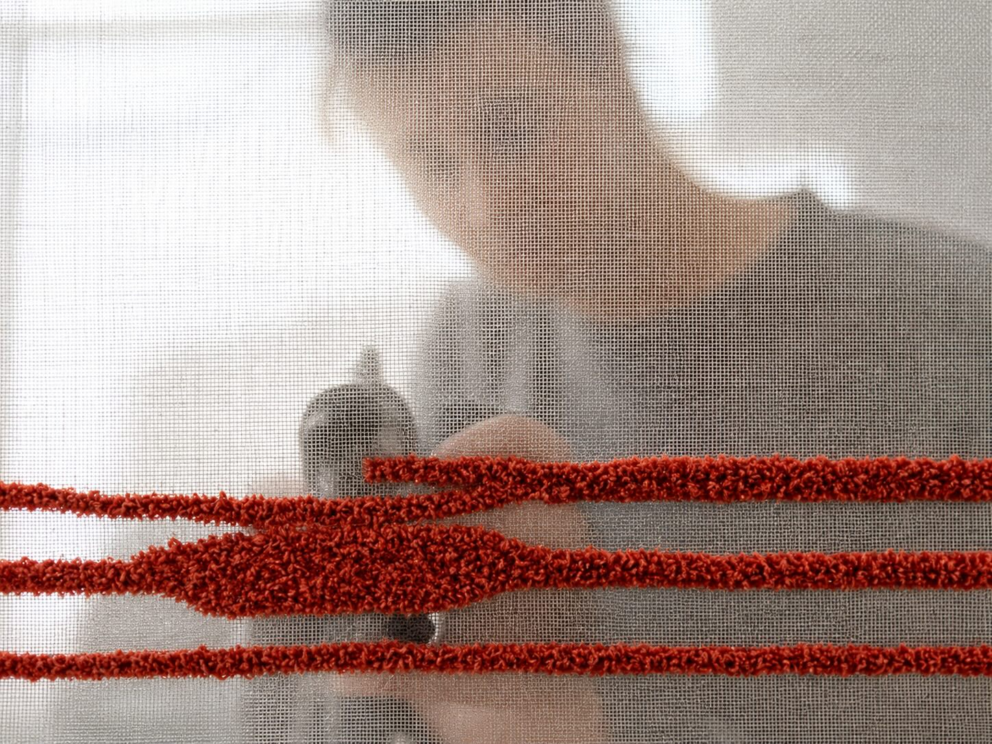 Close-up of a hand-tufted rug in production — red wool yarn punched through a stretched canvas backing with the artisan visible behind the mesh