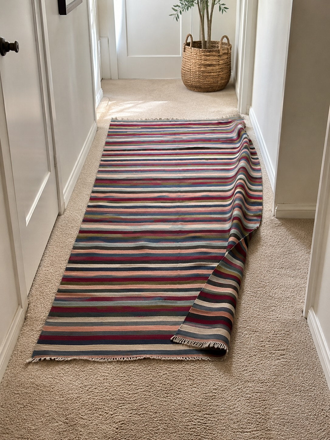 A vintage striped flatweave kilim runner rug visibly bunched and shifted on beige wall-to-wall carpet in a narrow hallway, showing how lightweight runner rugs slide without a rug pad