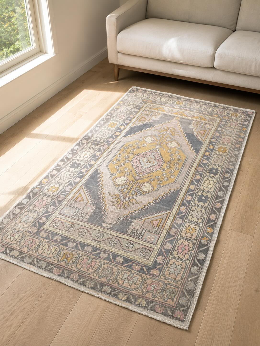 Vintage Turkish Oushak rug with faded central medallion in muted gray, gold and pink tones in a small room with light hardwood floor, neutral colors reflecting morning light and large-scale pattern keeping the eye moving inside the rug rather than toward the walls