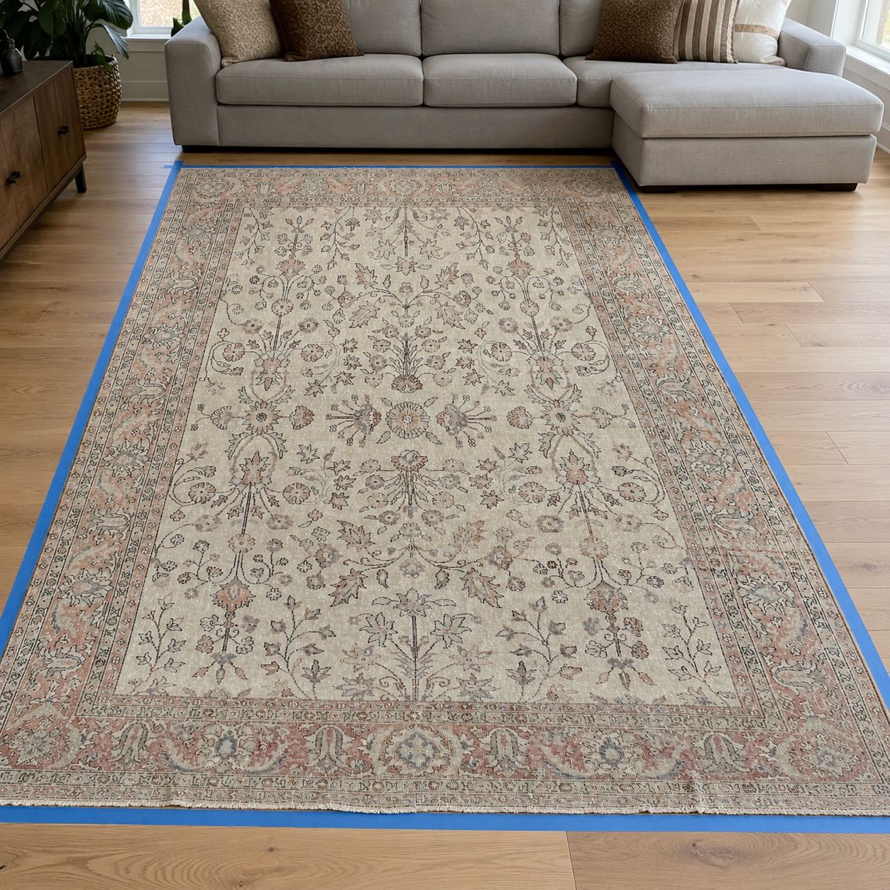 Blue painter's tape marking a 9x12 rug footprint on hardwood floor in front of a sectional sofa, showing how to visualize rug size before buying