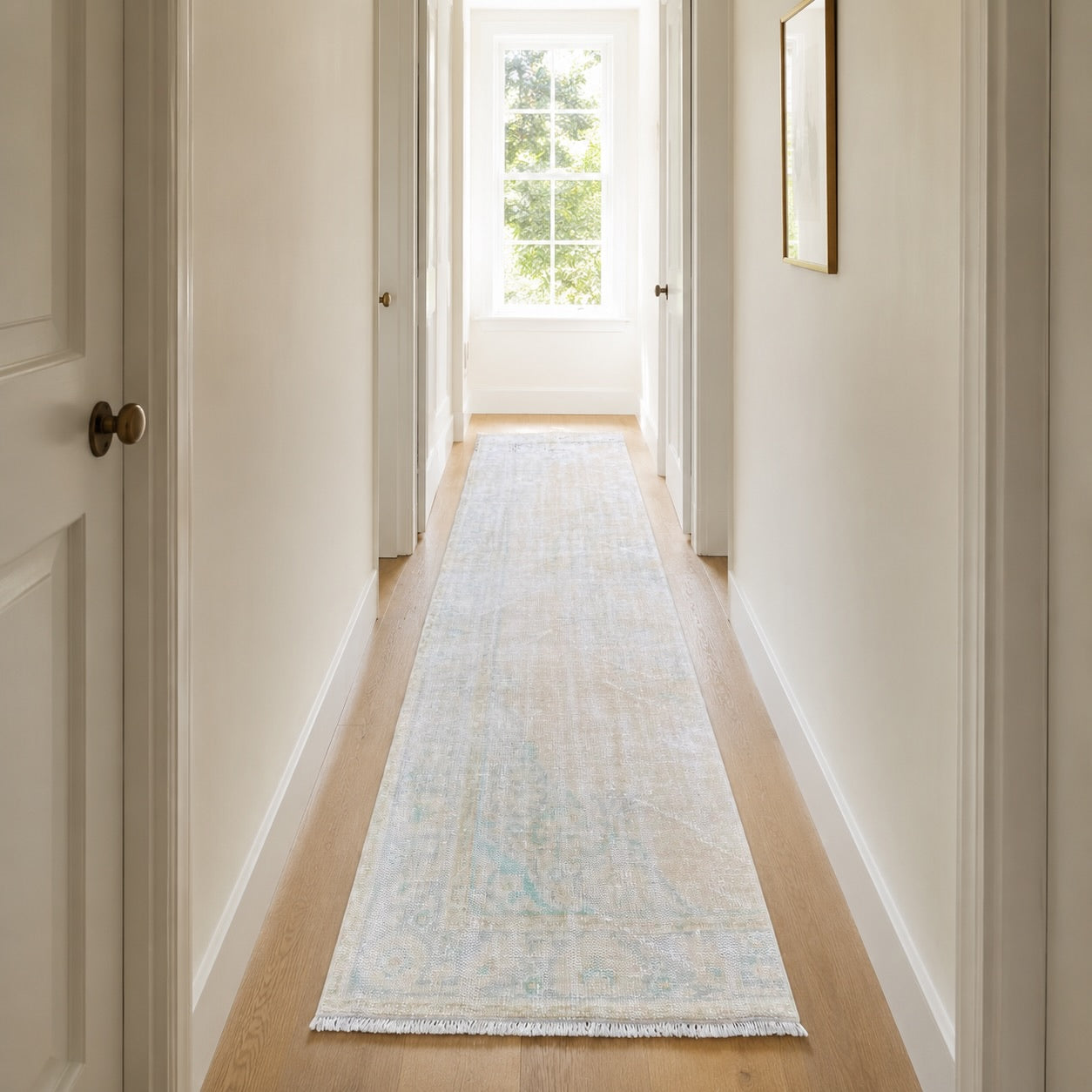 Faded vintage runner rug running the full length of a narrow white hallway with light oak floor, natural daylight from window at far end drawing the eye forward and making the corridor feel longer and more open