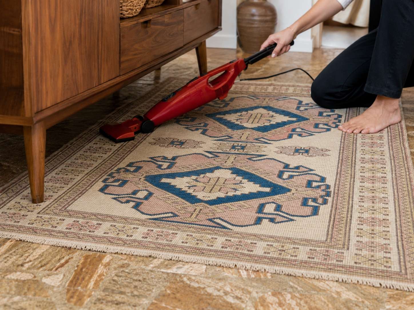 Red handheld vacuum cleaning under a mid-century sideboard on a vintage Turkish rug to remove carpet beetle larvae