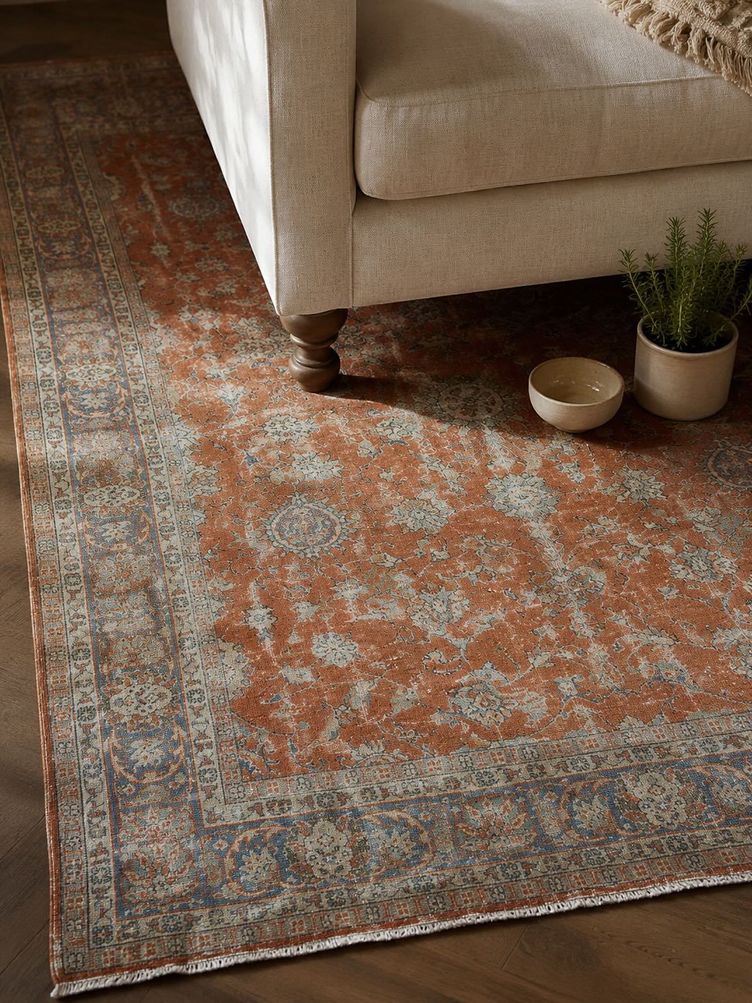 Close-up of a handwoven vintage Turkish rug in terracotta and blue tones beneath a linen sectional sofa leg, showing natural wool pile texture and floral Anatolian pattern